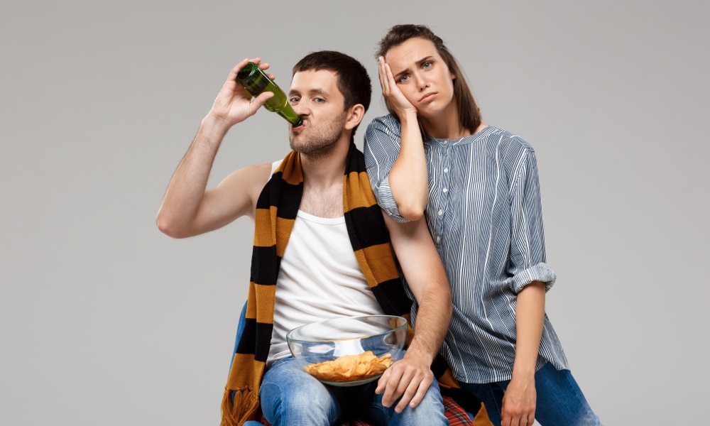 A man drinking a bottle of beer with chips on lap and woman leaning on him, exasperated.