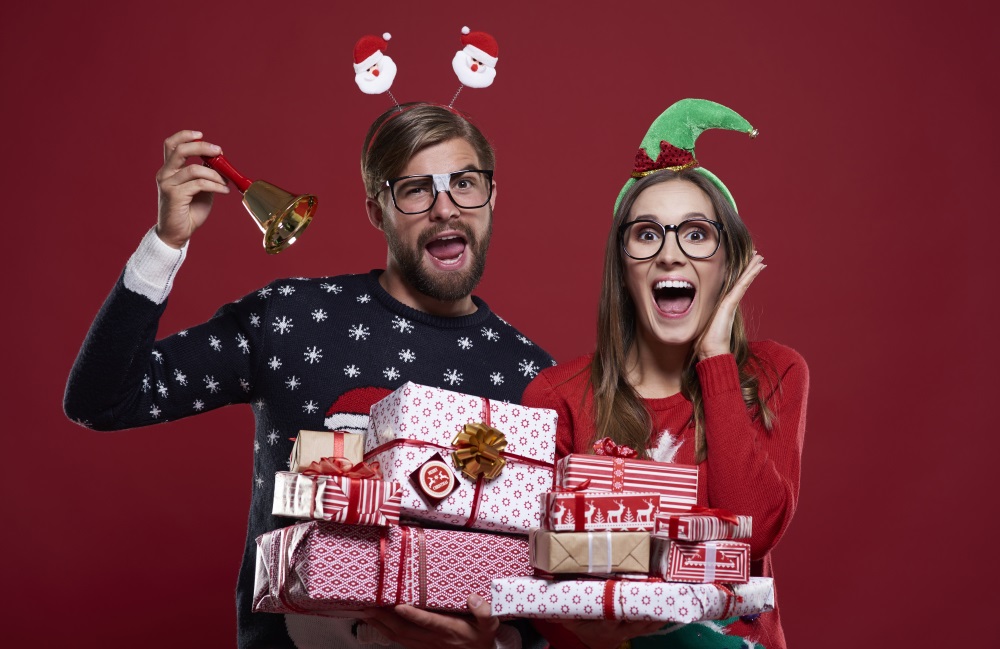 A goofy man and woman in Christmas clothing holding presents and making silly faces.