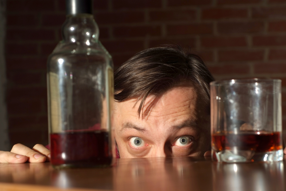 The top of a man's head, wide-eyed, staring at a bottle and glass on a bar or table.