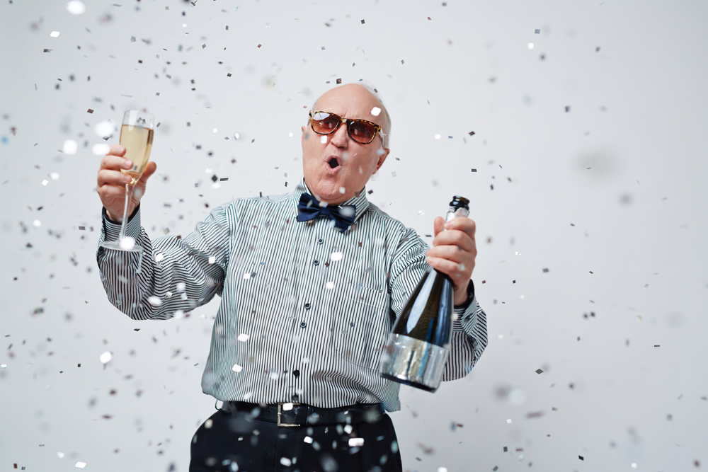 An older man wearing sunglasses holding a bottle of champaign and a glass