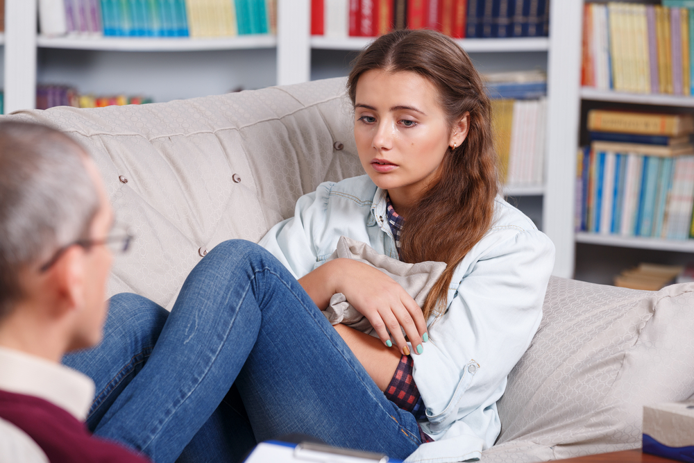 A crying young woman sits on a couch with her male therapist facing her.