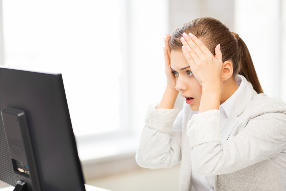 A woman looking at a computer screen, holding hands to her head, shocked or surprised.