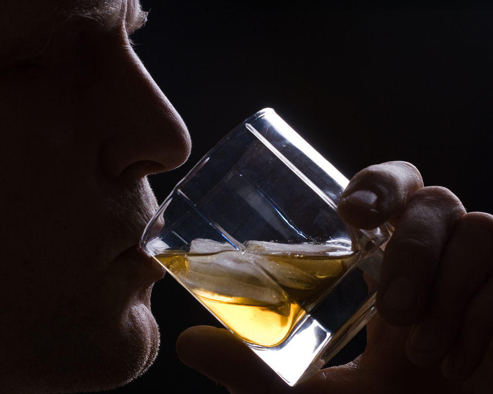 Man drinking whisky with ice from a glass against a dark background.