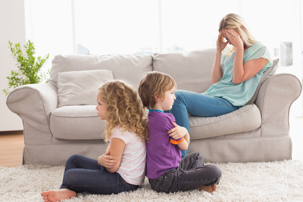 An upset mother on a couch with 2 small angry kids on the floor.