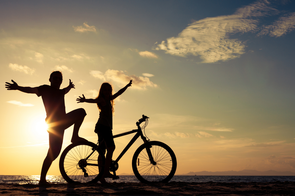 Father and daughter with bicycle, arms raised, against sunset.