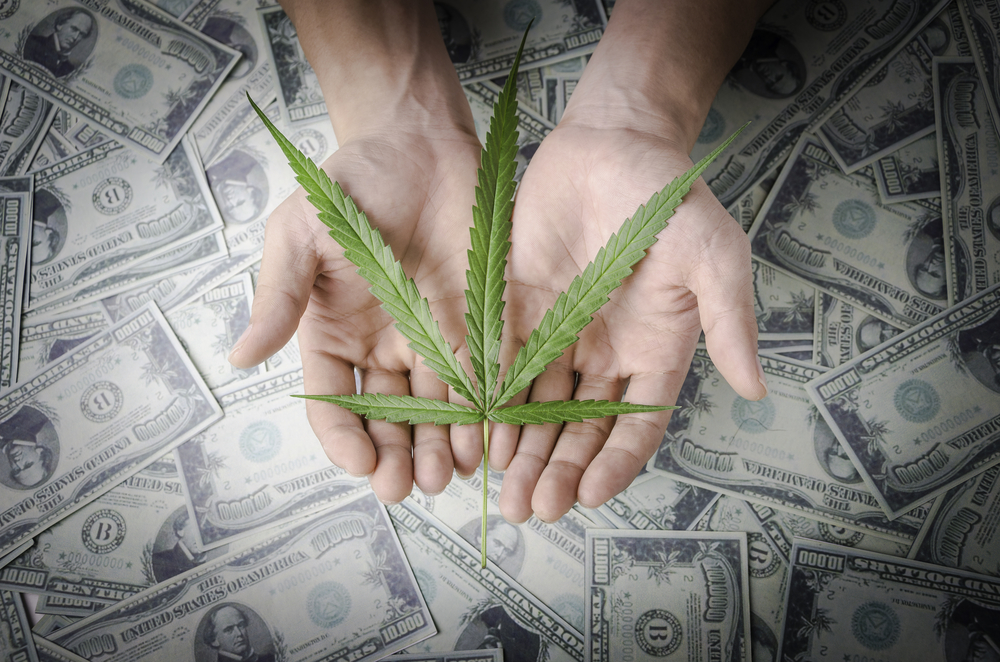 Hand holding a marijuana leaf floating above a pile of money.