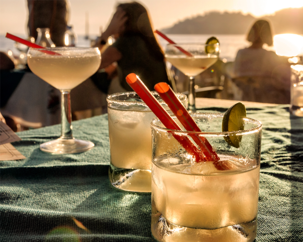 a variety of cocktails sitting on a table with a view of the beach