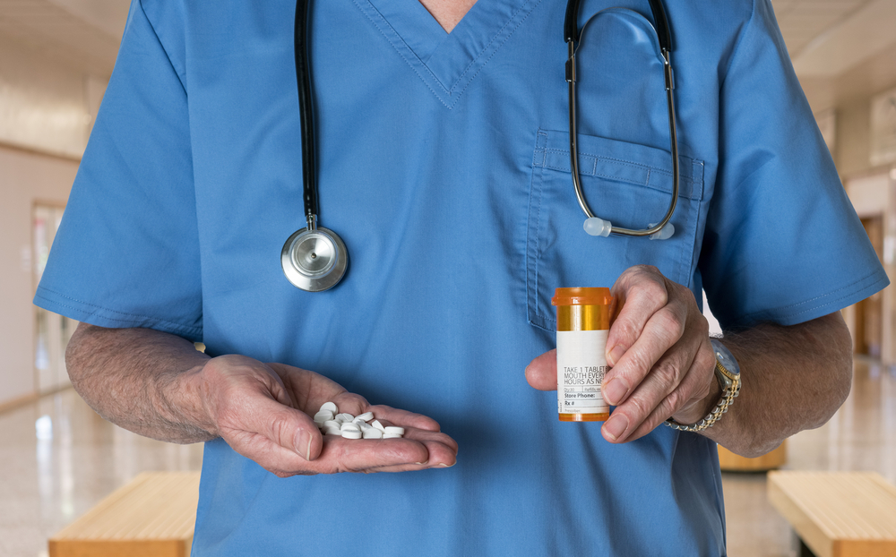 male doctor with stethoscope in medical scrubs holding a bottle and tablets of generic white RX tablets