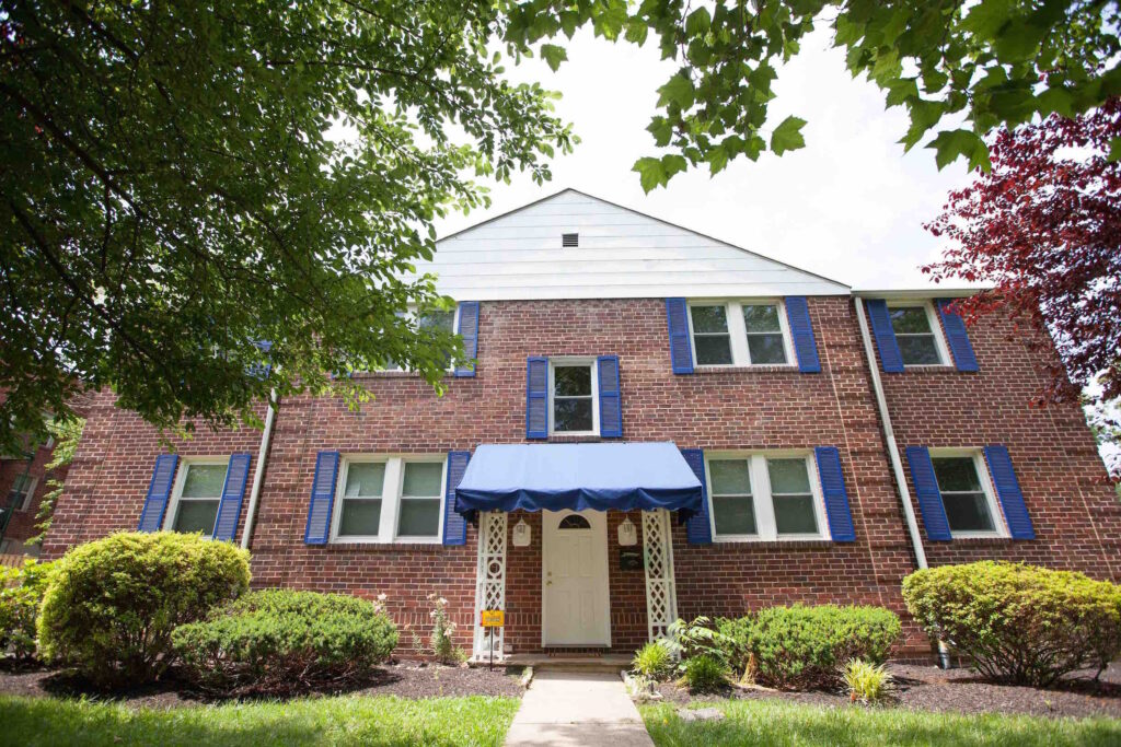 The front of a large brick house with blue awning and shutters, front yard with grass and trees.