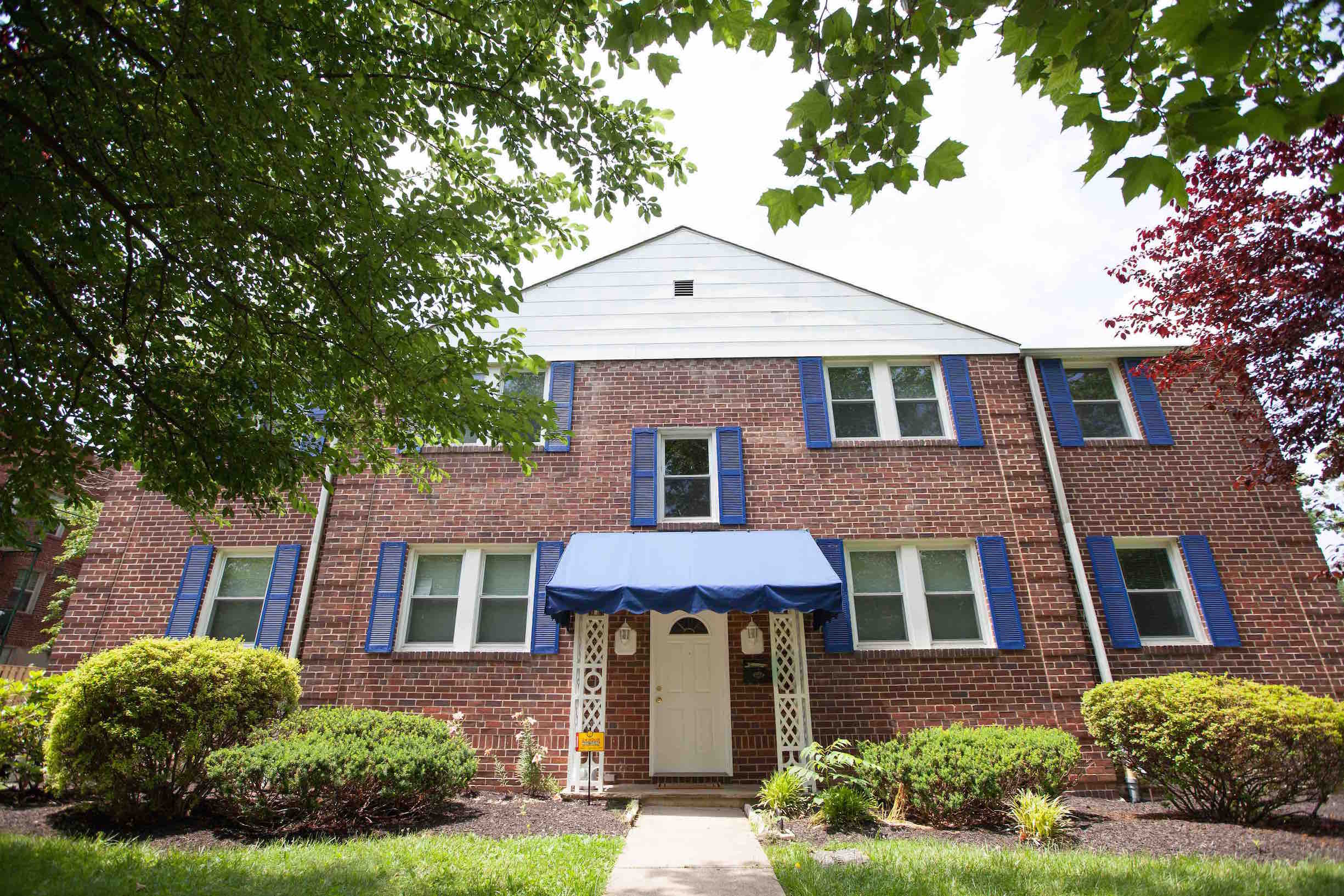 The front of a large brick house with blue awning and shutters, front yard with grass and trees.