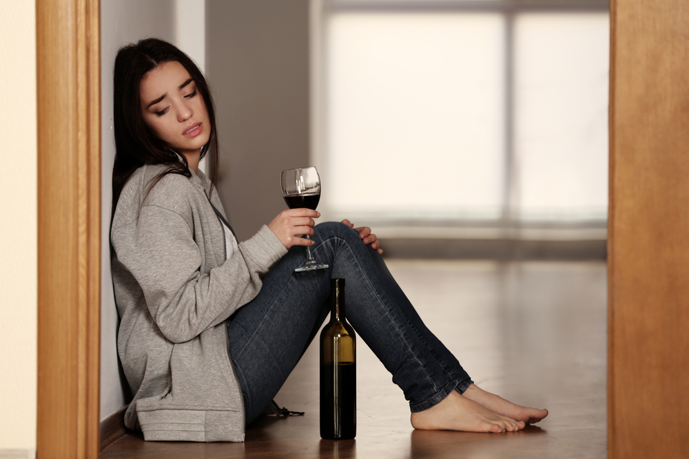 Woman on floor holding glass of wine, looking with concern at bottle of wine.