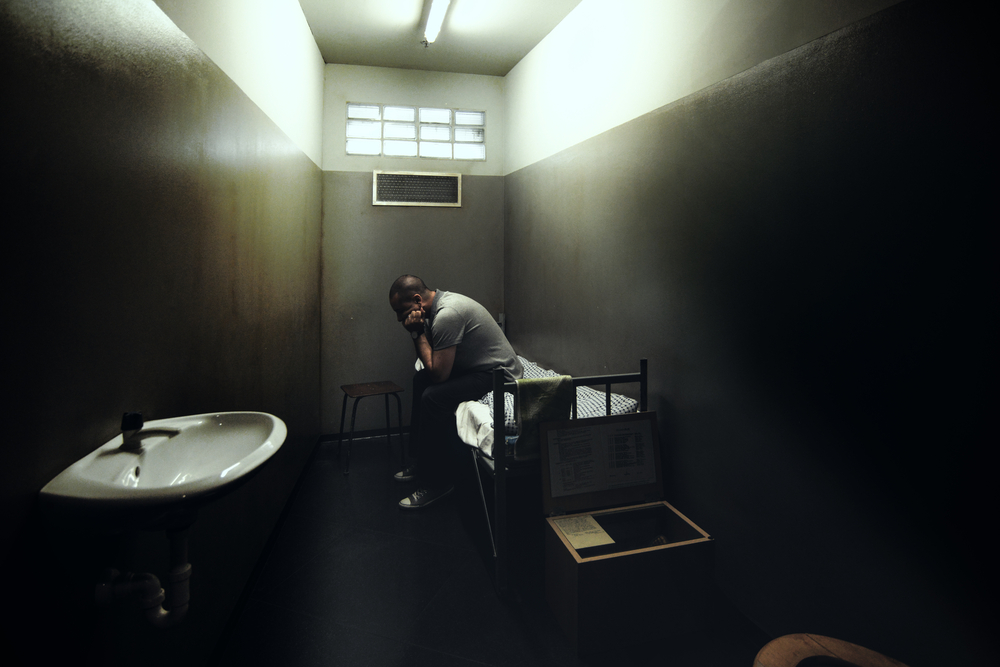One man sitting on a bed in a small room of a dark prison.