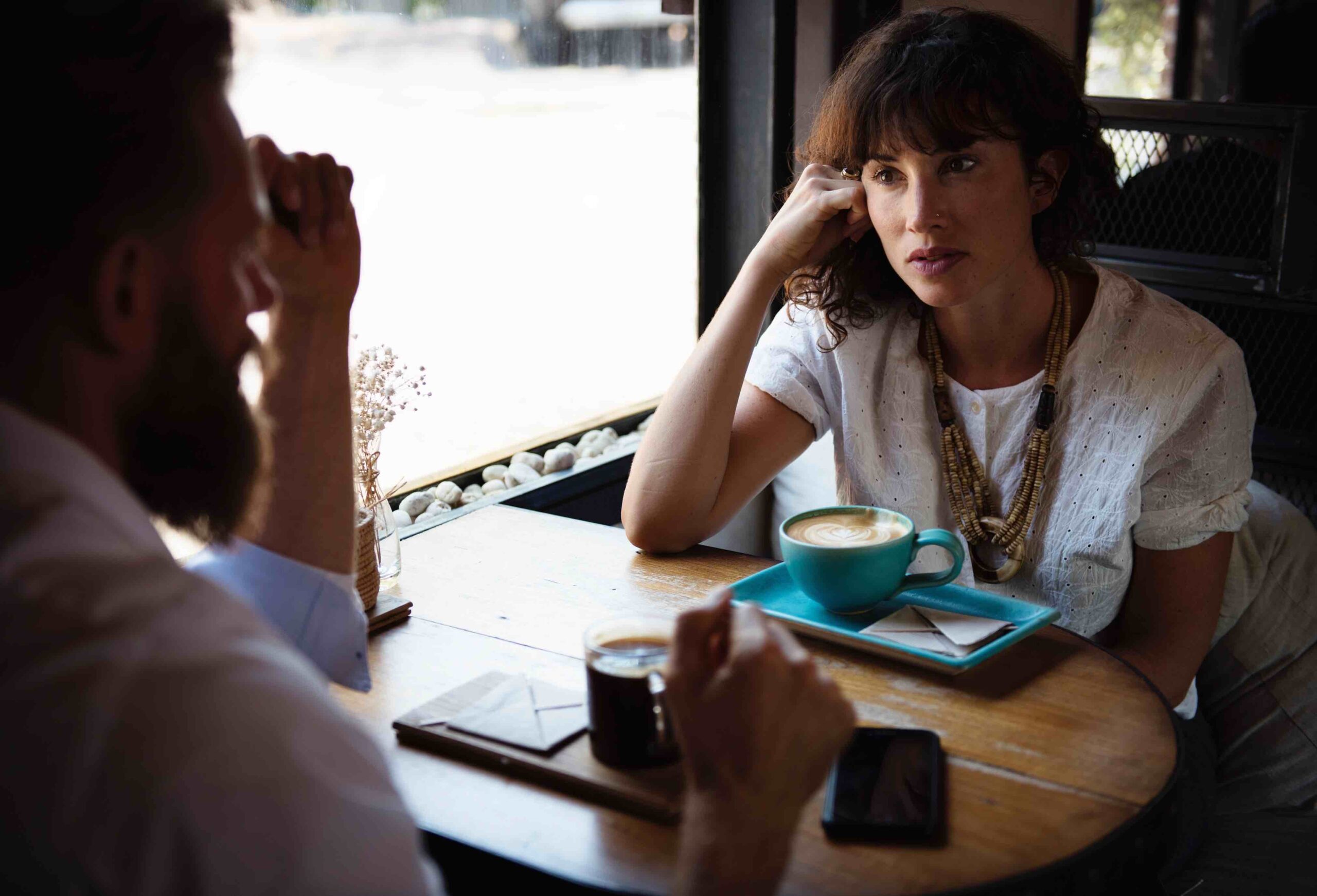 A woman sits at a table with coffee, head on hand, across from a man.