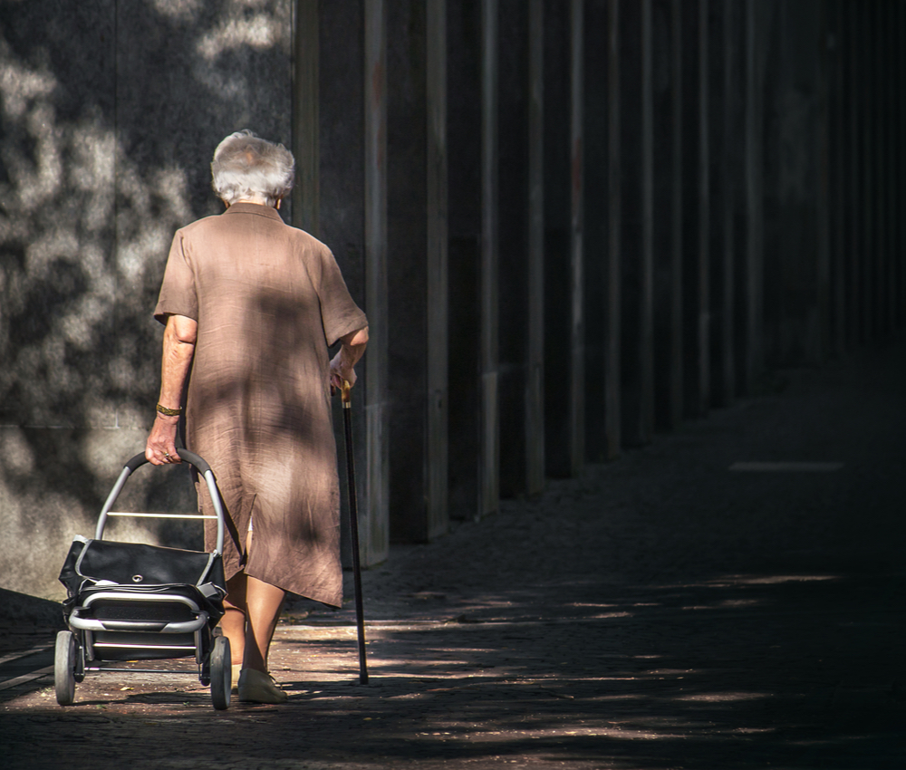 elderly woman walking alone with cane and suitcase with wheels going into the darkness