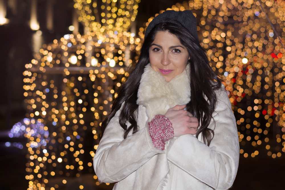 A woman in a white coat, in front of holiday lights.