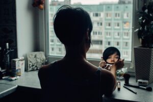 Woman sitting at a window, looking at her face in a compact mirror.