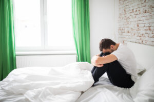 A man sitting on a bed with his knees pulled up, head down.
