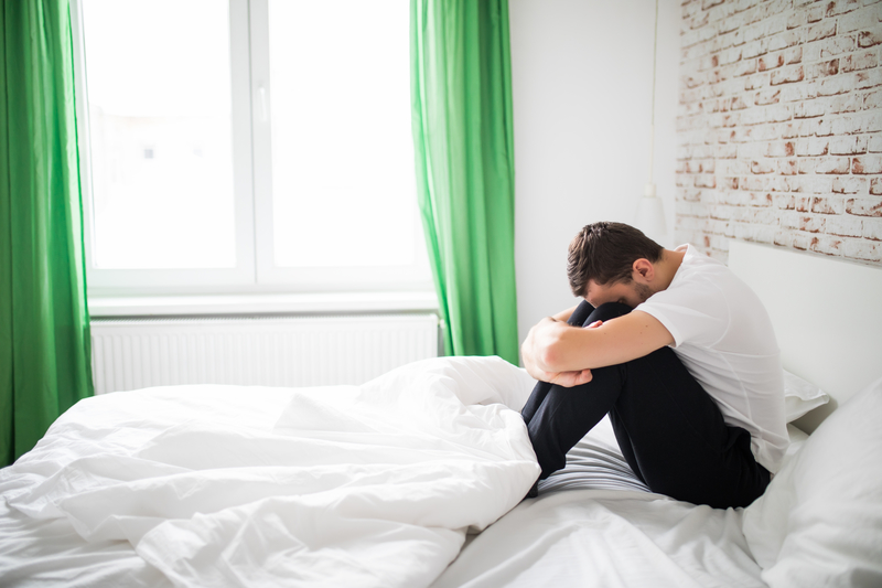 A man sitting on a bed with his knees pulled up, head down.