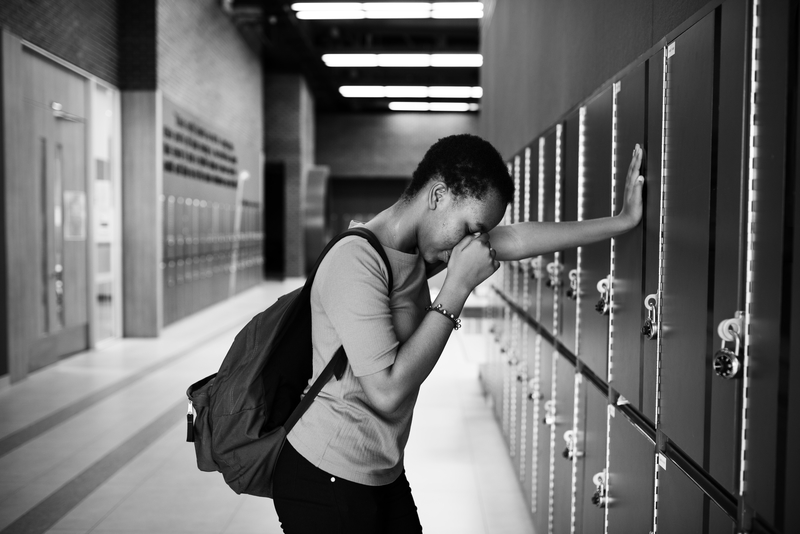 Black and white image of depressed woman student with backpack, leaning with one hand against lockers.