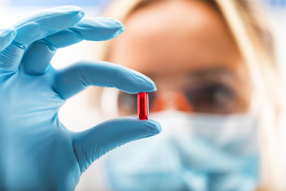 female scientist with protective eyeglasses and mask holding a red transparent pill with fingers in gloves