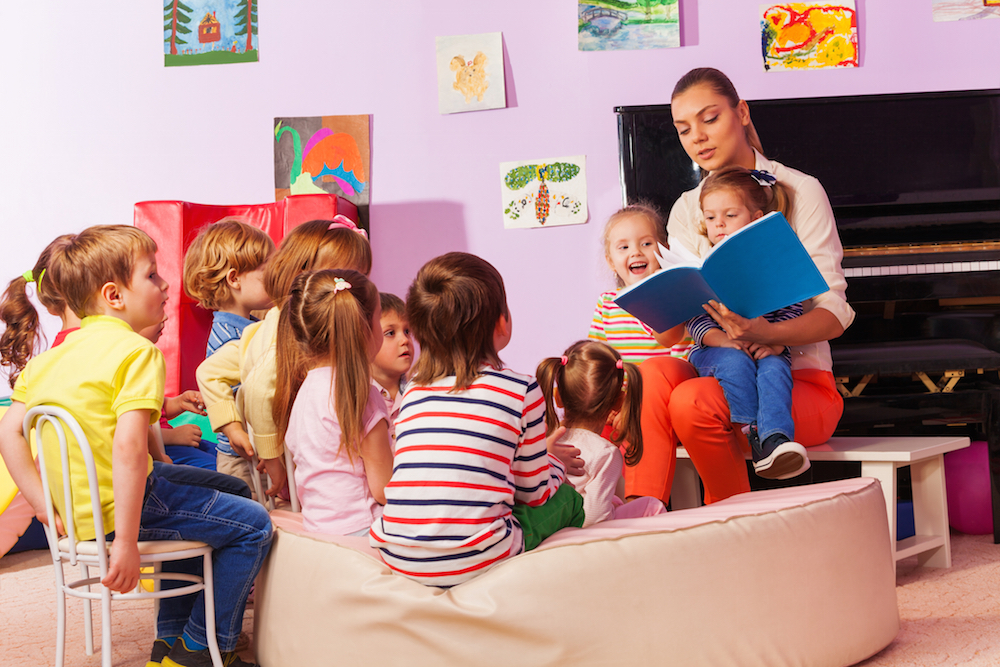 Large group of kids sit and listen to teacher reading a book and telling stories