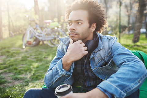 Thoughtful man sitting on the grass in a park with coffee, hand on chin.