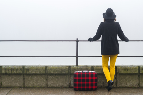 Back view of a woman standing at a railing with a suitcase next to her.