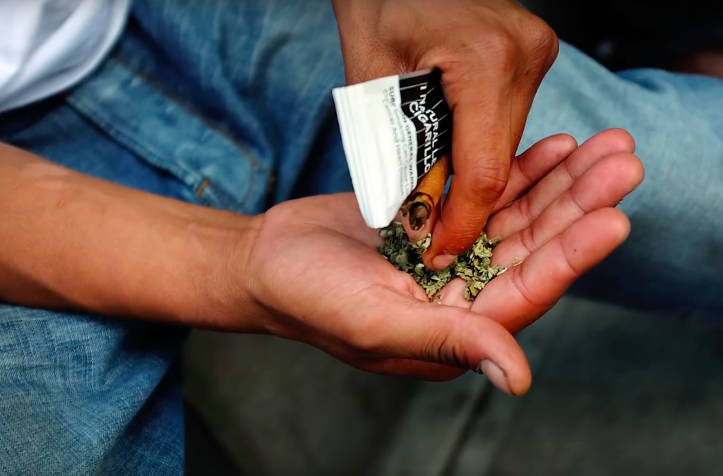 man holding a cigarillo and a handful of synthetic marijuana