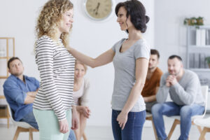 Two women standing together, smiling at each other, in front of a small group in a circle.