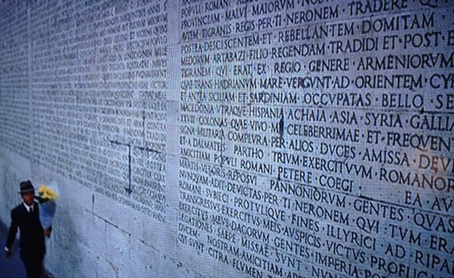 Man holding flowers walks by giant wall of text in Latin.