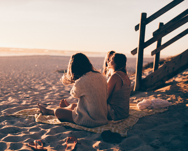 3 women site on a blanket on the sand.