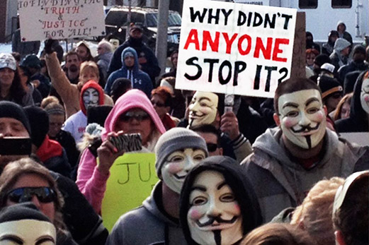 A crowd of people, some in Anonymous masks, holding sign reading "Why Didn't Anyone Stop It?"