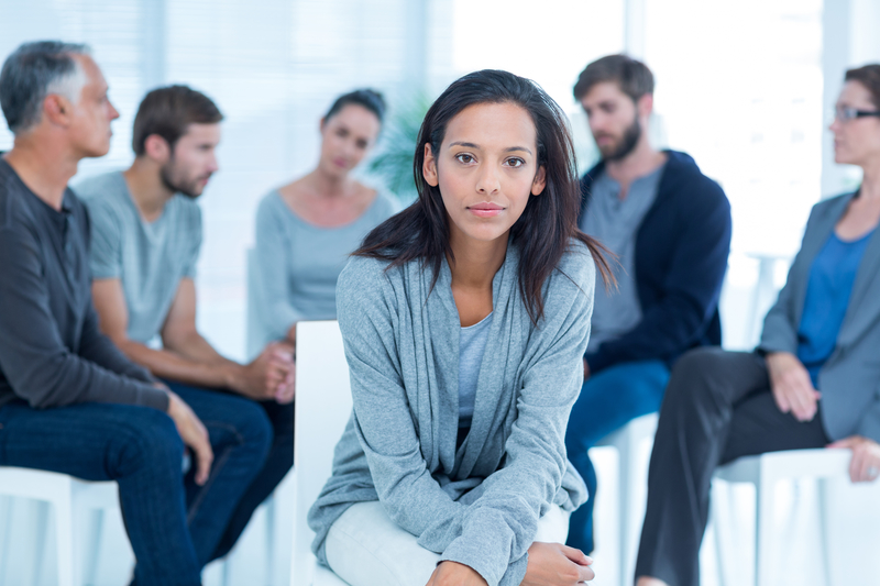 A group of people sit in a circle with one woman separate and facing the camera; group therapy
