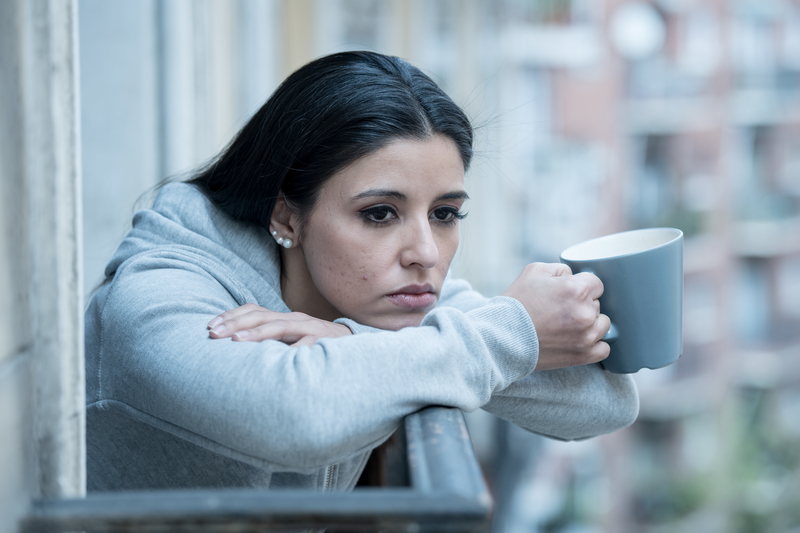 Woman holding coffee mug and resting chin on arms on a balcony; looks sad.