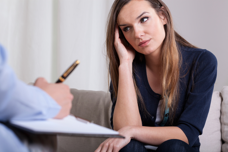 Woman sits with hand at temple, serious, facing someone who is writing on a notepad.