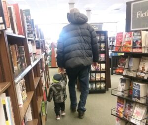 In a bookstore, a grown man and a toddler, holding hands, walk away from the camera.