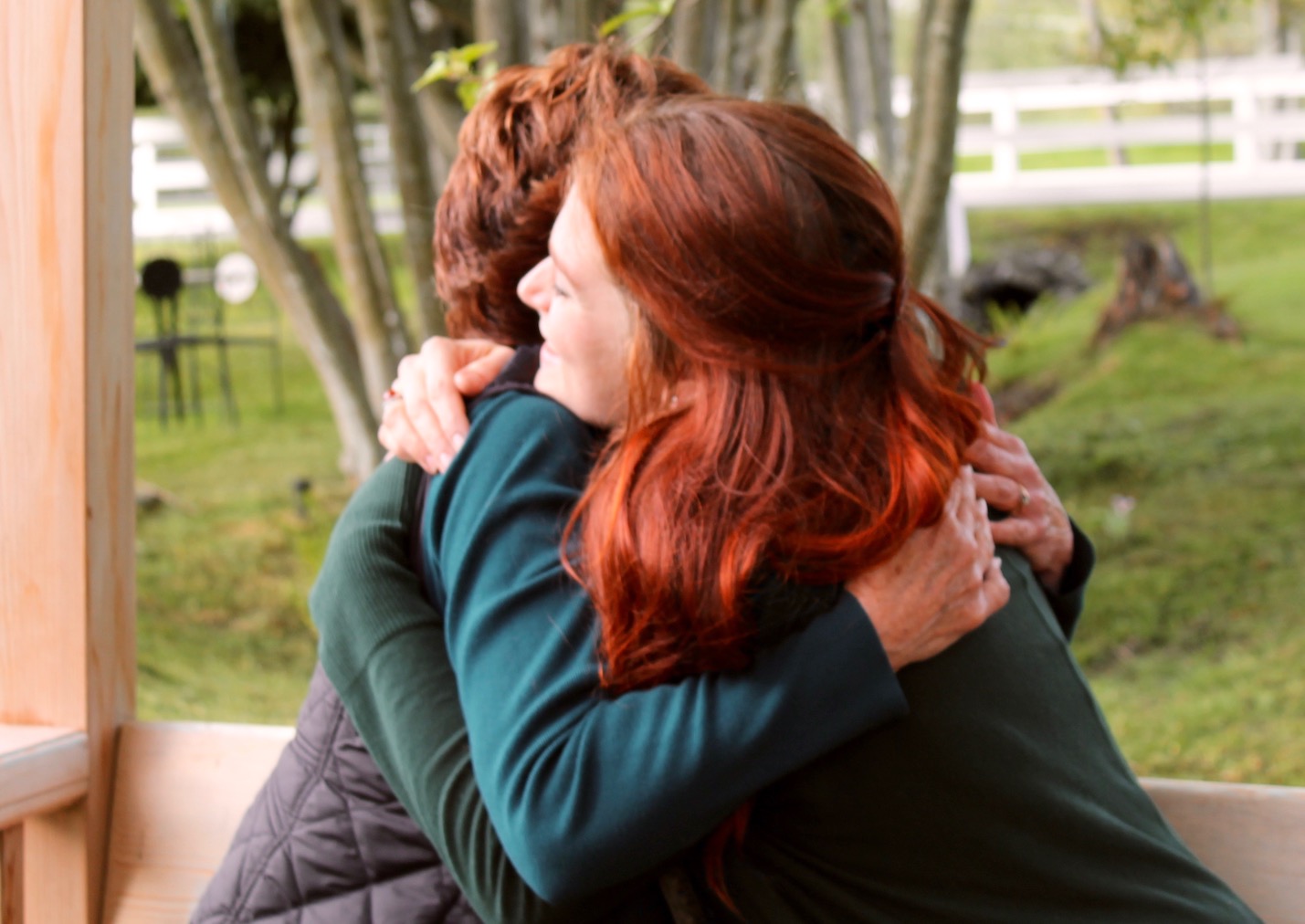 Two women embrace on what looks like a deck with green grass and white fence in the background.