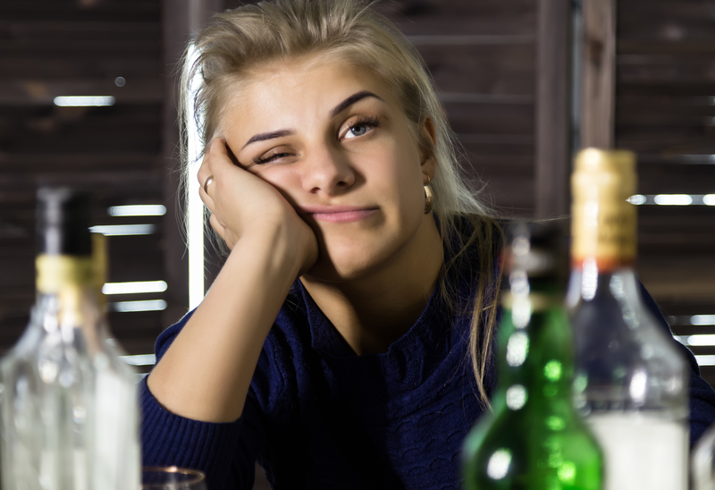 Woman with elbow on bar, face resting on hand, sits among alcohol bottles,