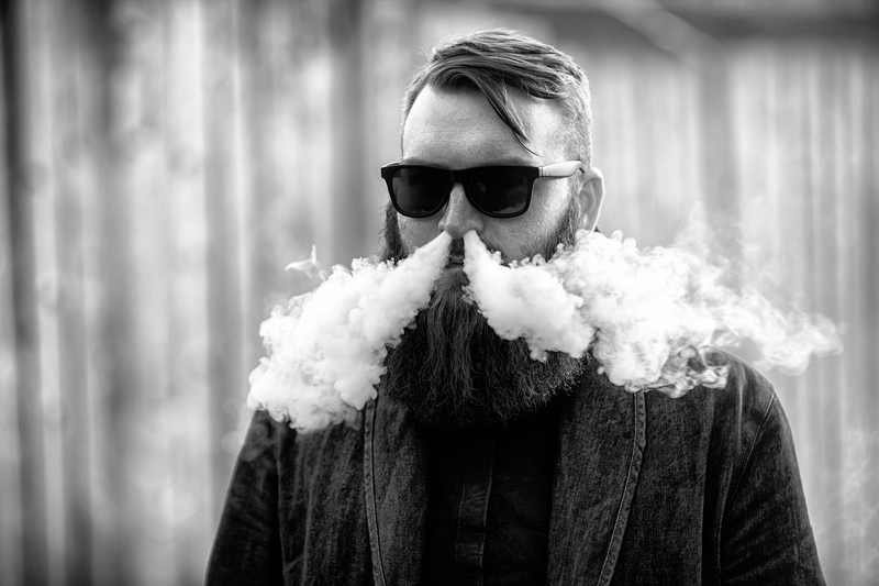 Vape man. Outdoor portrait of a young brutal white guy with large beard letting puffs out of steam from an electronic cigarette opposite the old wooden fence in the village. Vaping process. Black and white photo.