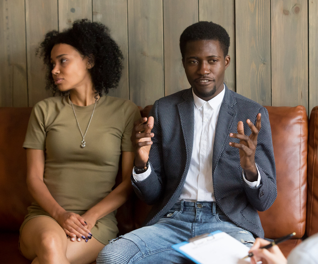 A couple sits on a therapist's couch. They appear to be in distress.