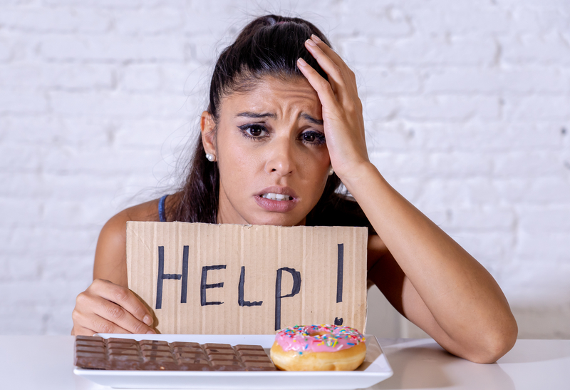 Woman with sugar addiction, has upset face and "Help!" sign in front of plate with sweets.