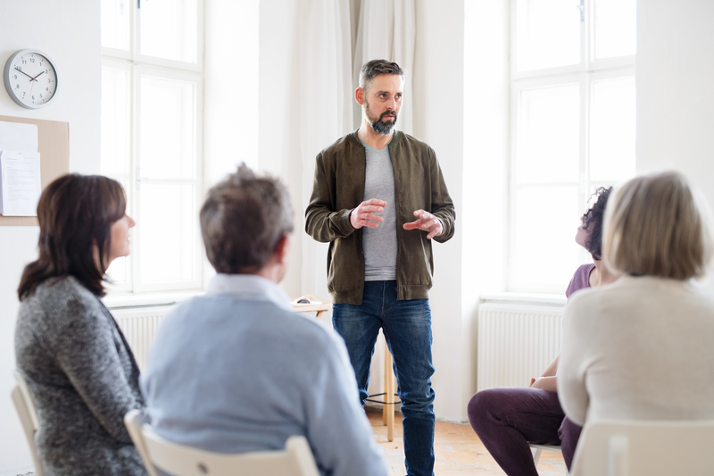 A group of people sit while one member stands, talking, AA meeting
