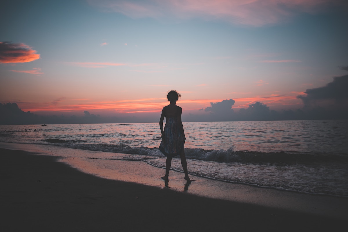 Woman at shoreline in front of setting sun, clouds, ocean