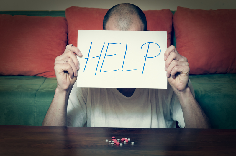 Man sitting in front of pile of pills holds sign that reads, "Help"