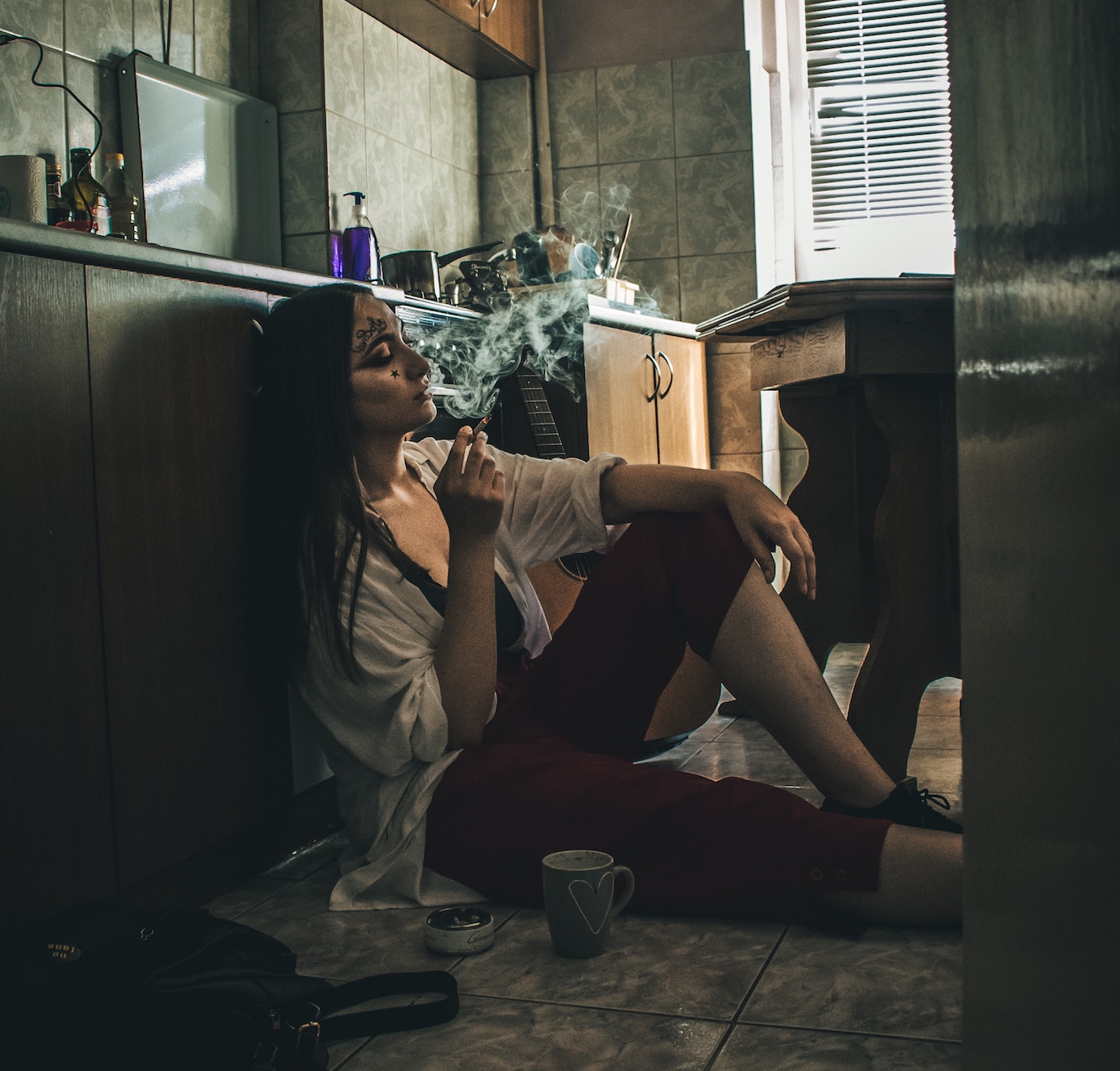 Woman sits on floor of kitchen, smoking a cigarette.