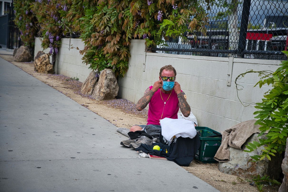Homeless man sits on ground wearing mask.