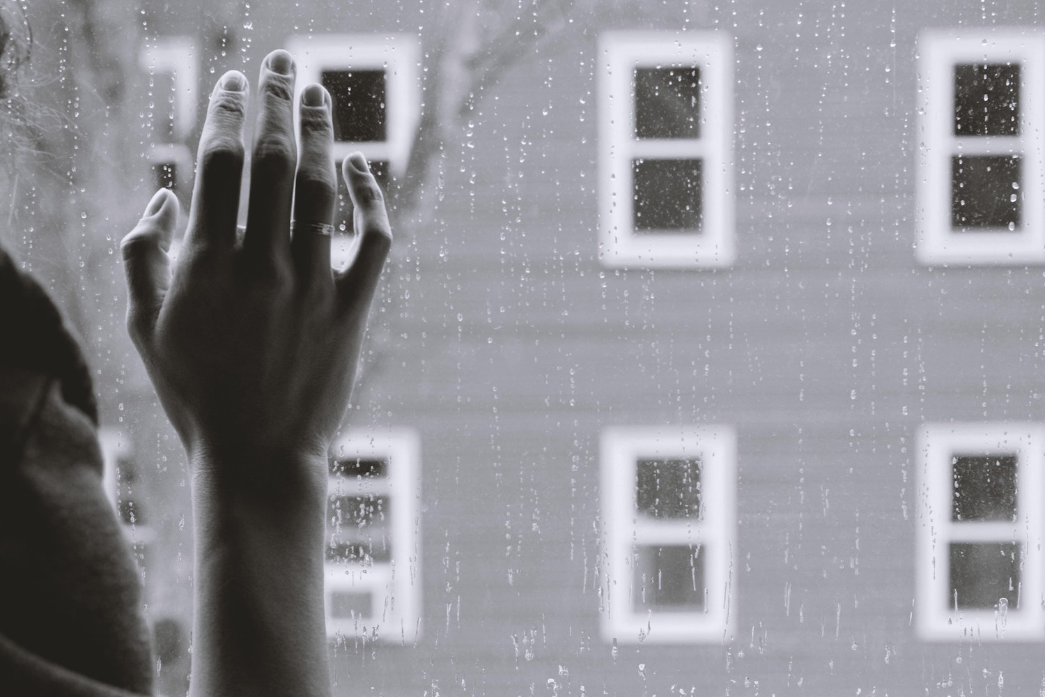 Black and white image, hand at window, rain and building outside.