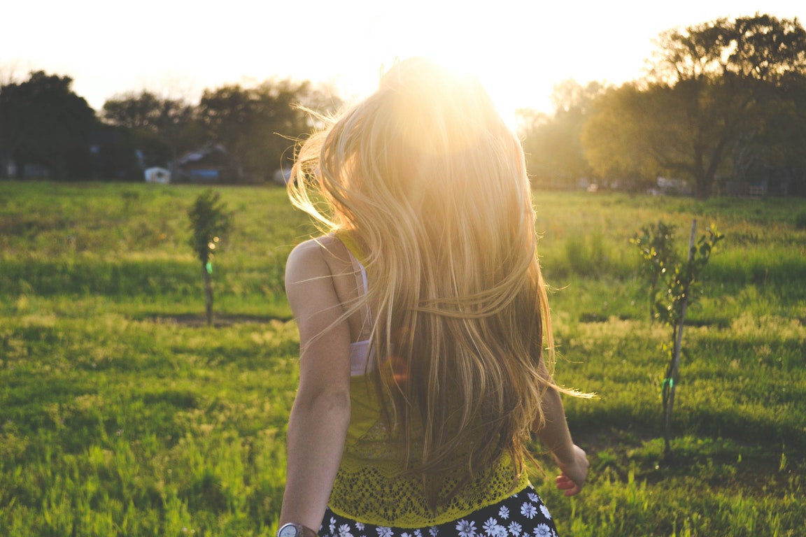 Girl with blonde hair walks away in a field, into sunshine