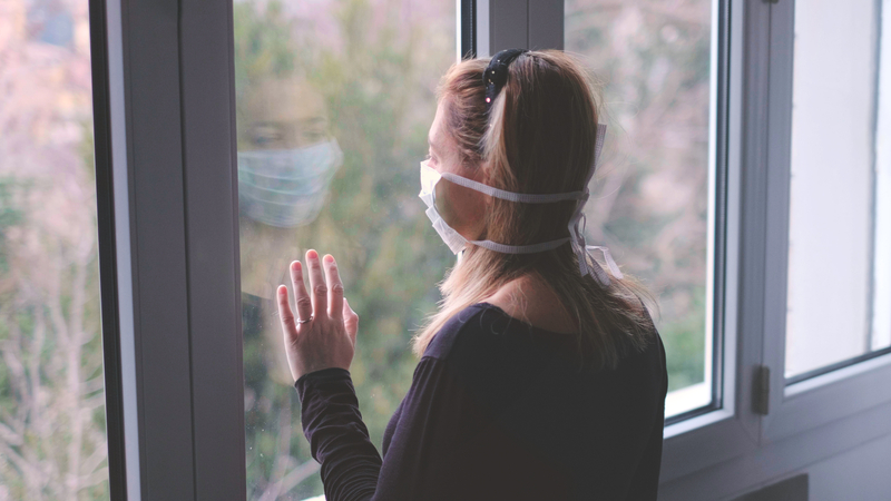 Woman with mask standing at window, hands on the pane.