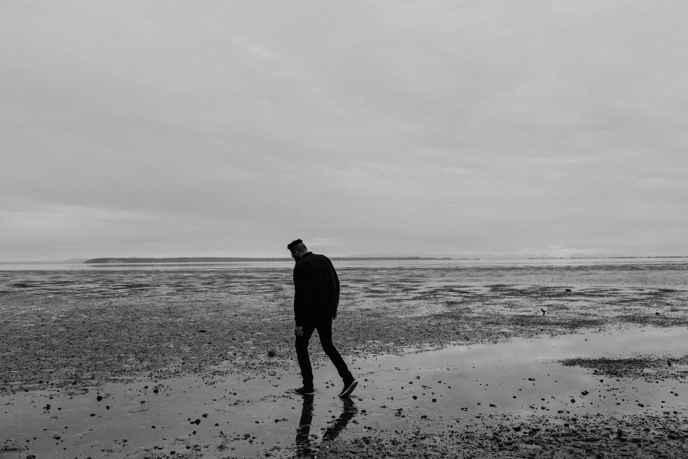 Black and white image of man walking alone on a rocky shore.